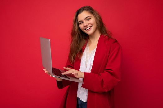 side profile photo of charming smiling confident pretty young lady holding laptop typing text on keyboard wearing red cardigan and white t-shirt isolated on red wall background.