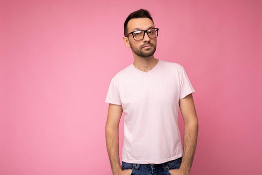 Handsome young self-confident brunet unshaven man wearing white t-shirt for mockup and stylish optical glasses isolated over pink background wall looking at camera. copy space