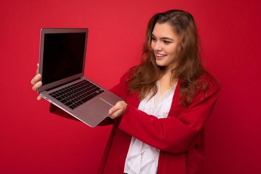 Photo of Beautiful smiling happy young brunette woman holding computer laptop with empty monitor screen wearing red cardigan and white blouse looking at netbook isolated over red background.