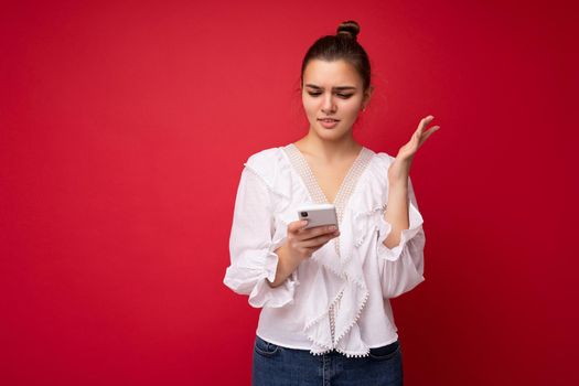 Photo shot of attractive dissatisfied sad good looking young woman wearing casual stylish outfit poising isolated on background with empty space holding in hand and using mobile phone messaging sms looking at smartphone display screen.