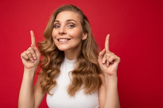 Photo of attractive positive surprised smiling young woman pointing fingers up at copyspace presenting ads promo with wow omg sincere emotions wearing good look outfit isolated over background with free space.