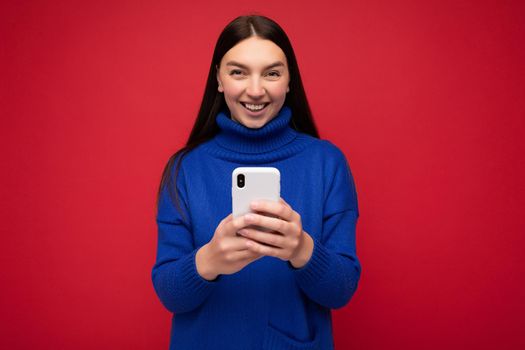 Smiling attractive positive good looking young brunette woman wearing stylish blue warm sweater poising isolated on red background with empty space holding in hand and using mobile phone messaging sms looking at camera.