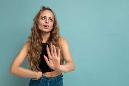 Young tired upset attractive winsome blonde curly woman with sincere emotions wearing trendy black top isolated over blue background with free space and showing stop gesture saying no. Negative concept.