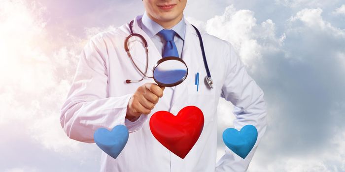 Horizontal shot of confident doctor in white medical suit is looking at heart symbols through magnifier while standing against cloudy skyscape view on background.