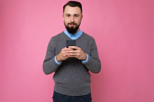 handsome good looking brunet bearded young man wearing grey sweater and blue shirt isolated on pink background with empty space holding in hand and using mobile phone communicating online looking at camera.