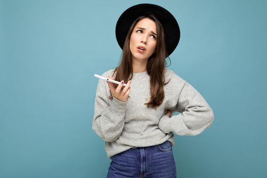 Beautiful young brunette woman thinking wearing black hat and grey sweater holding smartphone looking up texting isolated on background.Copy space