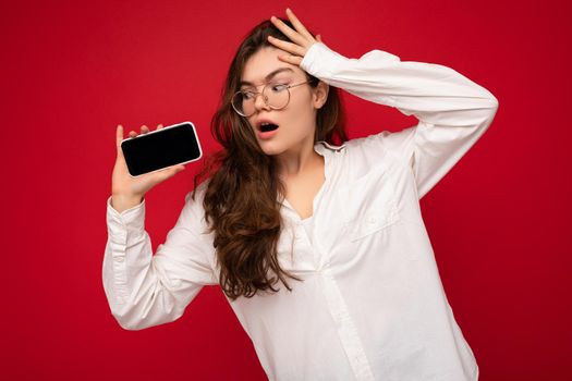 Amazed charming young brunet woman wearing white shirt and optical glasses isolated over red background holding in hand and showing mobile phone with empty screen for mockup looking at gadjet screen.