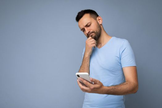 Photo shot of handsome thinking good looking young man wearing casual stylish outfit poising isolated on background with empty space holding in hand and using mobile phone messaging sms looking at smartphone display screen.