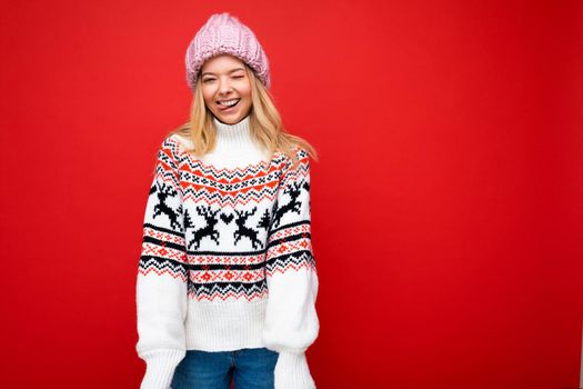 Photo of pretty positive young blonde woman isolated over red background wall wearing winter sweater and pink hat looking at camera. Copy space