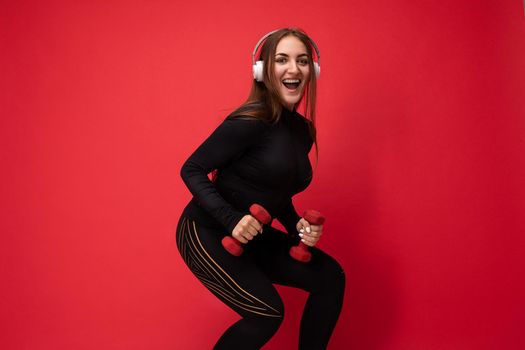 Side-profile photo of beautiful happy smiling young brunet woman wearing black sport clothes isolated on red background wall squating using dumbbells wearing white headsets listening to music looking at camera.