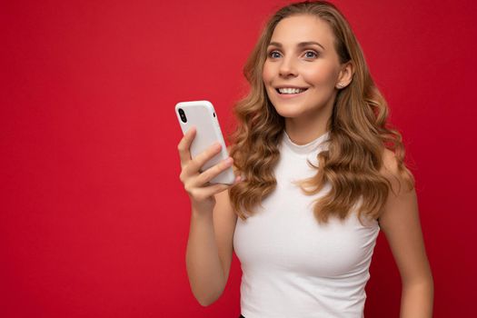closeup Smiling happy pretty young blonde woman wearing white t-shirt isolated over red background using smartphone and chatting via mobile phone.
