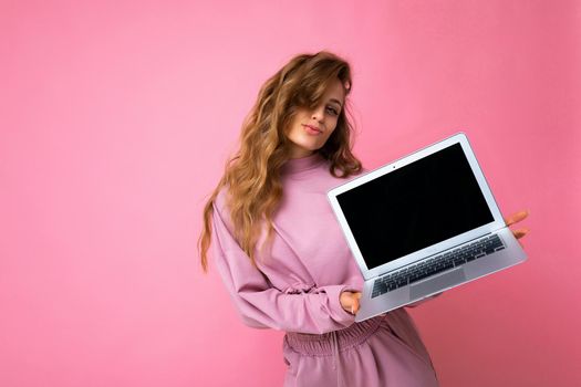Photo of beautiful blond curly woman holding computer laptop with empty monitor screen with mock up and copy space wearing pink sweater isolated over pink wall background.