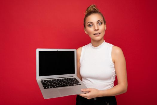 beautiful blond young woman looking at camera holding computer laptop wearing white t-shirt isolated over red wall background.