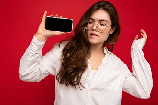 photo of sexy beautiful young brunette woman wearing white shirt and optical glasses isolated over red background holding in hand and showing mobile phone with empty screen for cutout looking at gadjet screen.