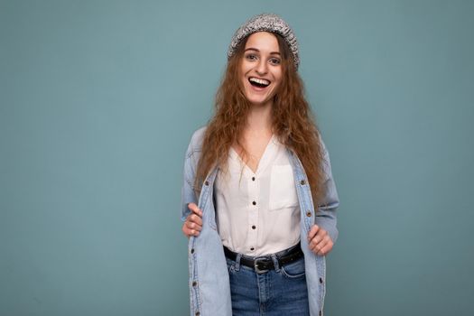 Photo of beautiful happy smiling young dark blonde curly woman standing isolated over blue background wall wearing jean shirt white shirt and grey hat and looking at camera. Copy space