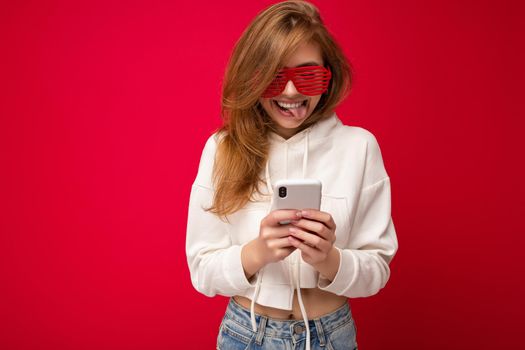 Photo shot of attractive positive good looking young woman wearing casual stylish outfit poising isolated on background with empty space holding in hand and using mobile phone messaging sms looking at smartphone display screen.