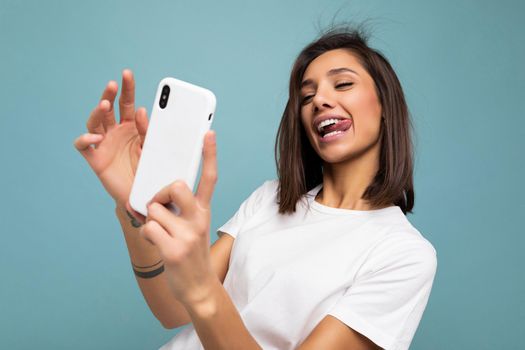 Photo of sexy beautiful smiling happy young brunette woman wearing casual white t-shirt isolated over wall blue background holding and using mobile phone taking selfie looking at gadjet screen and showing tongue.