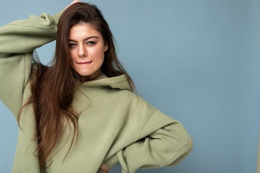 Portrait of beautiful young caucasian smiling brunette woman model in trendy khaki hoodie isolated on blue background with copy space. Positive concept.