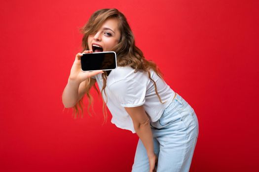 Photo of beautiful smiling young blonde woman good looking wearing white t-shirt standing isolated on red background with copy space holding phone showing smartphone in hand with empty screen display for mockup looking at camera.