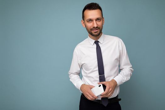 Photo of handsome good looking brunet unshaven man with beard wearing casual white shirt and tie isolated on blue background with empty space holding in hand mobile phone looking at camera. Copy space