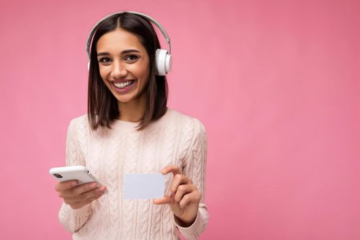 Photo shot of beautiful joyful smiling young female person wearing stylish casual outfit isolated over colorful background wall wearing white bluetooth wireless earphones and listening to music and using mobile phone and credit card making payment looking at camera.