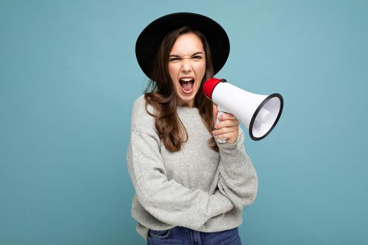 Photo of young beautiful positive brunette woman with sincere emotions wearing stylish black hat and grey sweater isolated over blue background with copy space and screaming in magaphone.