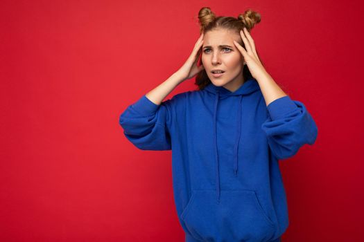 Photo of emotional shocked amazed young pretty blonde woman with two horns with sincere emotions wearing hipster bright blue hoodie isolated over red background with copy space.