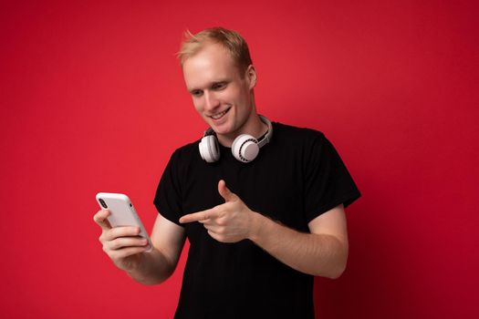 Photo shot of handsome positive good looking young man wearing casual stylish outfit poising isolated on background with empty space holding in hand and using mobile phone messaging sms looking at smartphone display screen.