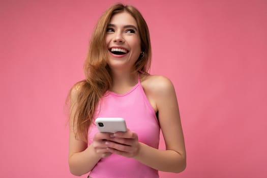 Photo of smiling happy good looking young blonde woman wearing pink top poising isolated on pink background with empty space holding in hand and using mobile phone messaging sms looking up and dreaming.