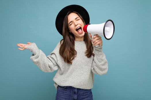 Photo of young beautiful positive emotional brunette woman with sincere emotions wearing stylish black hat and gray sweater isolated over blue background with copy space and shouting into magaphone.