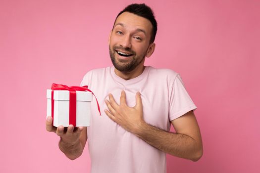 Photo shot of handsome positive msmiling brunette unshaven young man isolated over pink background wall wearing pink t-shirt holding white gift box with red ribbon and looking at camera.
