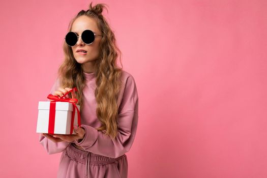 Shot of beautiful positive young blonde curly woman isolated over pink background wall wearing pink sport clothes and sunglasses holding gift box and unboxing surprise looking to the side. Empty space