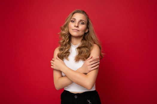 Photo of young charming fascinating attractive happy touchy blonde woman with sincere emotions wearing stylish clothes isolated over red background with copy space and embraces herself.