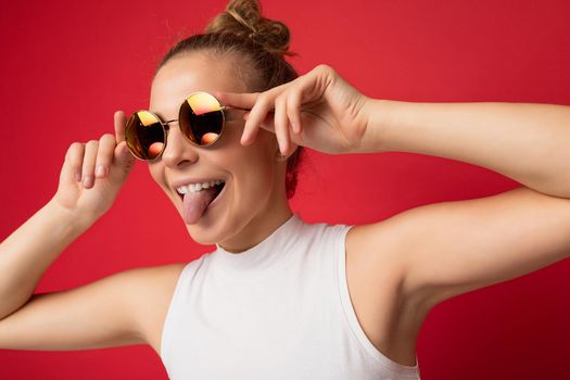 Closeup photo of attractive young blonde woman wearing everyday stylish clothes and modern sunglasses isolated on colorful background wall looking to the side and showing tongue.