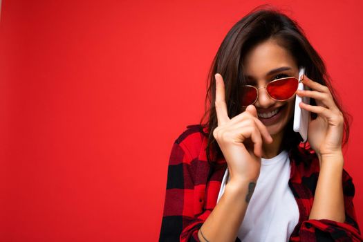 Closeup photo of sexy pretty happy young brunet woman wearing stylish red shirt white t-shirt and red sunglasses isolated over red background communicating on mobile phone looking at camera.