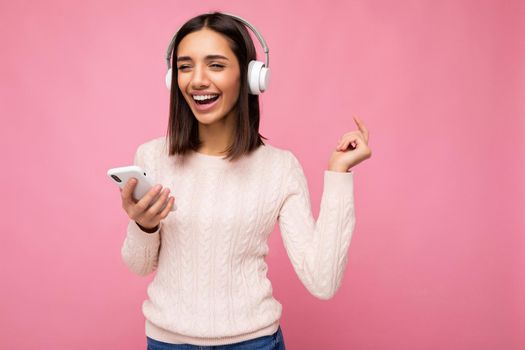 Photo of beautiful joyful smiling young woman wearing stylish casual clothes isolated over background wall holding and using mobile phone wearing white bluetooth headphones listening to music and having fun looking to the side.