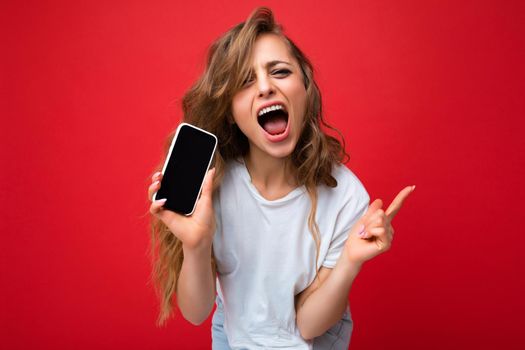 Photo of beautiful joyful smiling young blonde woman good looking wearing white t-shirt standing isolated on red background with copy space holding phone showing smartphone in hand with empty screen display for mockup looking at camera and shouting.