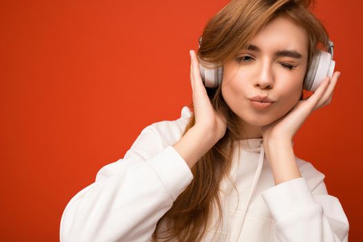 Closeup photo of attractive cute positive young blonde woman wearing white hoodie isolated over colourful background wearing white wireless bluetooth headphones listening to good music looking at camera and giving kiss.