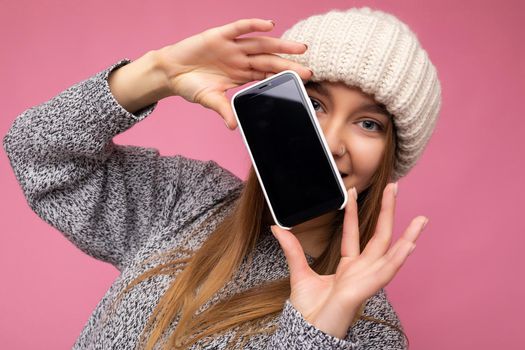 Photo of beautiful smiling young woman good looking wearing casual stylish outfit standing isolated on background with copy space holding smartphone showing phone in hand with empty screen display for mockup looking at camera.