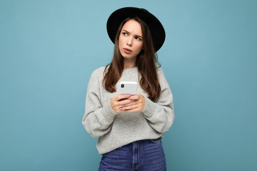 Beautiful young brunette woman thinking wearing black hat and grey sweater holding smartphone looking to the side texting isolated on background.Copy space