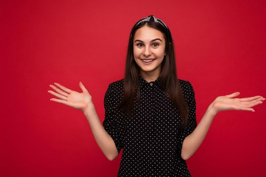 Photo of beautiful smiling positive brunette girl standing isolated over red wall wearing casual stylish black clothes looking at camera and showing two open palms.