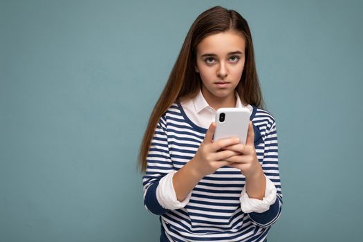 Photo shot of attractive upset sad good looking teenager wearing casual stylish outfit poising isolated on background with empty space holding in hand and using mobile phone messaging sms looking at camera.