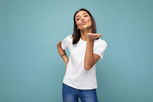 Photo of positive young beautiful brunette woman with sincere emotions wearing casual white t-shirt for mockup isolated over blue background with copy space and giving kiss.