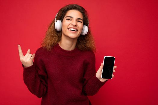 Shot of attractive positive smiling young brunet curly woman wearing dark red sweater isolated on red background wall wearing white wireless headphones and listening to music and showing mobile phone with empty screen for cutout looking at camera and showing rock and roll sign.