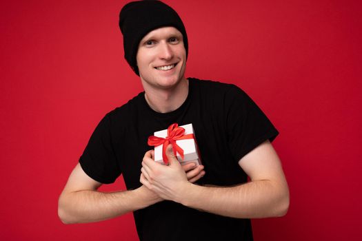 Shot of handsome positive smiling young man isolated over red background wall wearing black hat and black t-shirt holding white gift box with red ribbon and looking at camera.