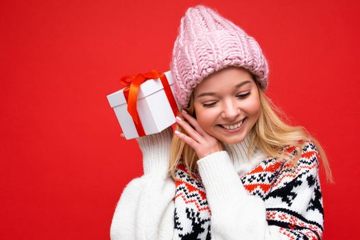 Attractive positive smiling young blonde woman isolated over red background wall wearing winter sweater and pink hat holding white gift box with red ribbon and looking down.