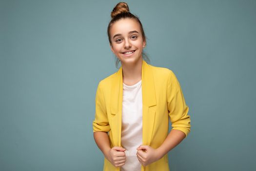 Shot of beautiful happy smiling brunette little girl wearing trendy yellow jacket and white t-shirt standing isolated over blue background wall looking at camera. Empty space