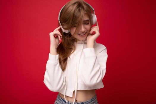 Attractive emotional positive young blonde woman wearing white hoodie isolated over colourful background wearing white wireless bluetooth headsets listening to music and moving.