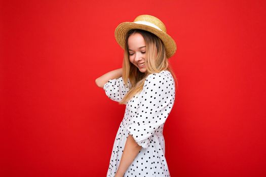 Portrait of young beautiful smiling hipster blonde woman in trendy summer dress and straw hat. Sexy carefree female person posing isolated near red wall in studio. Positive model with natural makeup. Copy space.