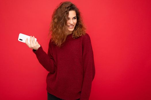 Photo of pretty smiling happy young woman with curly hair wearing dark red sweater isolated on red background holding mobile phone looking to the side and having fun.